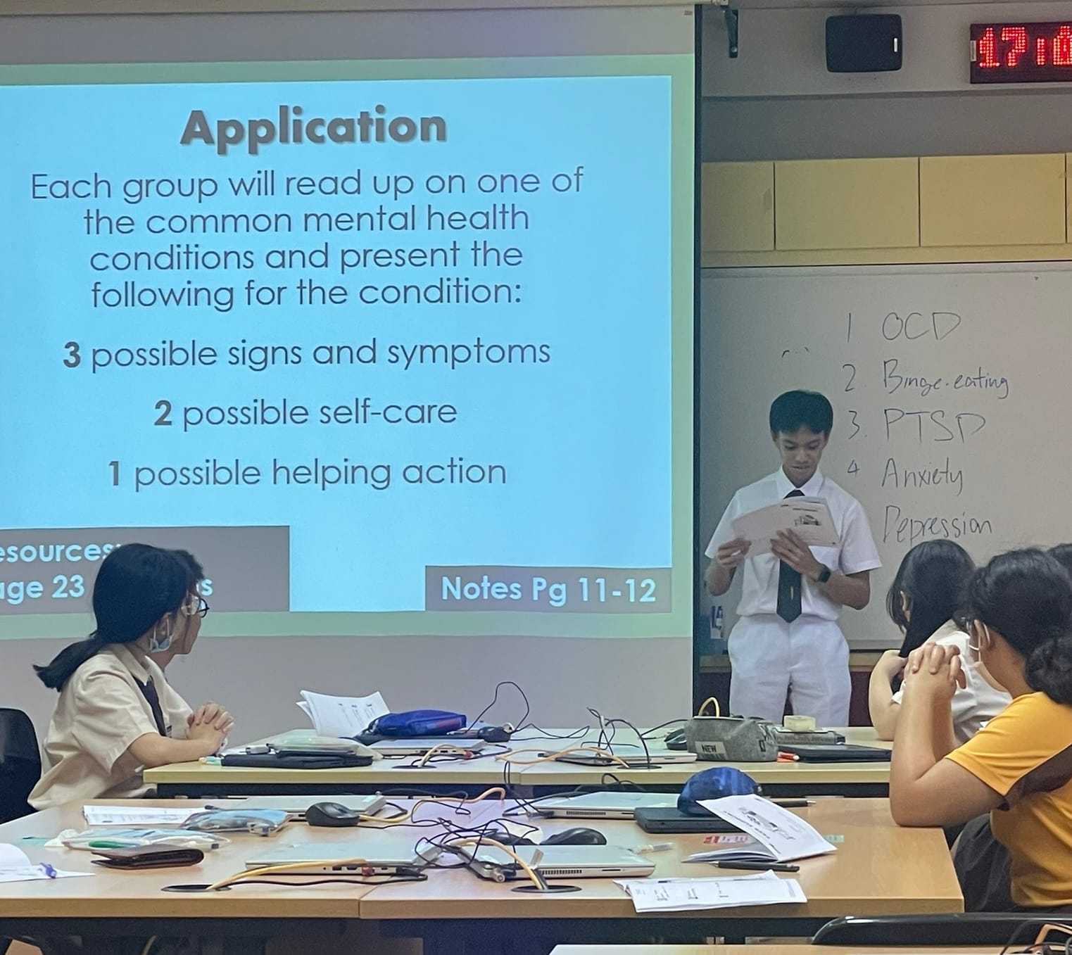 A student presents in a classroom beside a slide on mental health conditions and a whiteboard listing OCD, PTSD, and Anxiety.